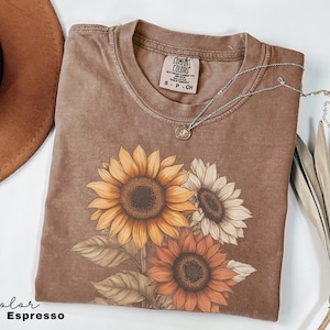 May include: A brown t-shirt with a faded look features a graphic of three sunflowers in shades of yellow, orange, and white. The text "COMFORT COLORS" is visible on the shirt's neckline.