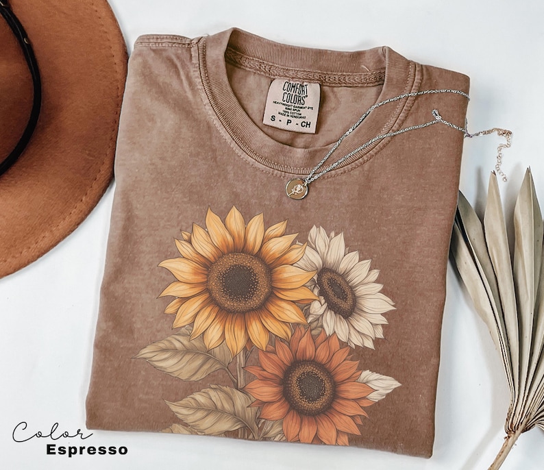 May include: A brown t-shirt with a faded look features a graphic of three sunflowers in shades of yellow, orange, and white. The text "COMFORT COLORS" is visible on the shirt's neckline.