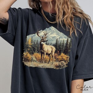 May include: Black t-shirt featuring a detailed graphic of a deer in a mountain landscape. The design includes a deer with large antlers, trees, and a mountain range. The words "Color Black" are written in cursive.