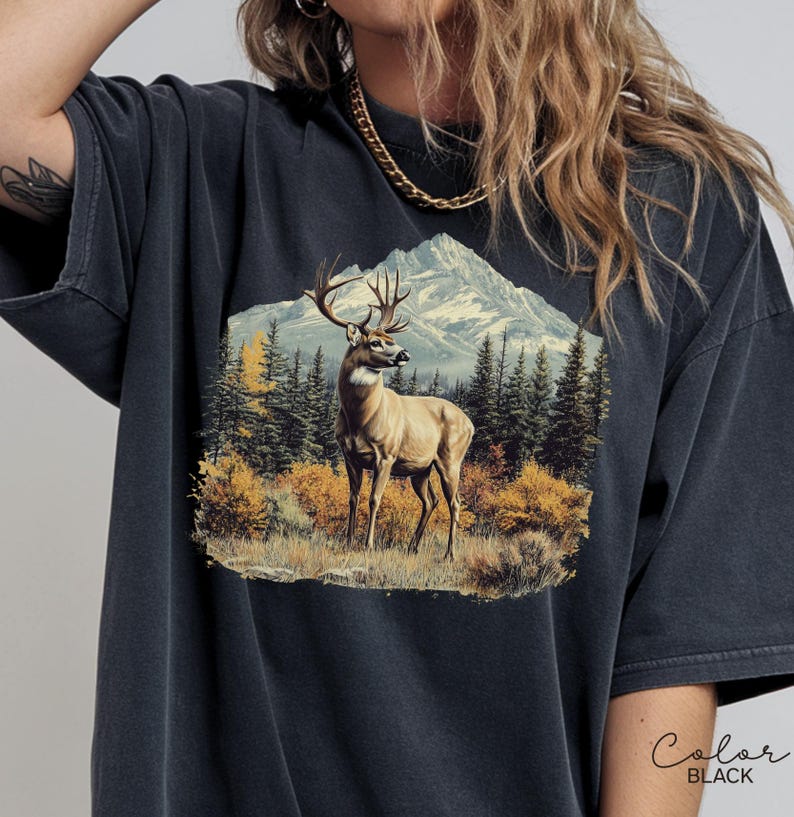 May include: Black t-shirt featuring a detailed graphic of a deer in a mountain landscape. The design includes a deer with large antlers, trees, and a mountain range. The words "Color Black" are written in cursive.