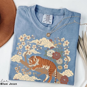 May include: Blue jean colored t-shirt with a tiger graphic. The tiger is resting on a tree branch with flowers and clouds. The shirt has a crew neck and short sleeves. A gold necklace is draped across the shirt.