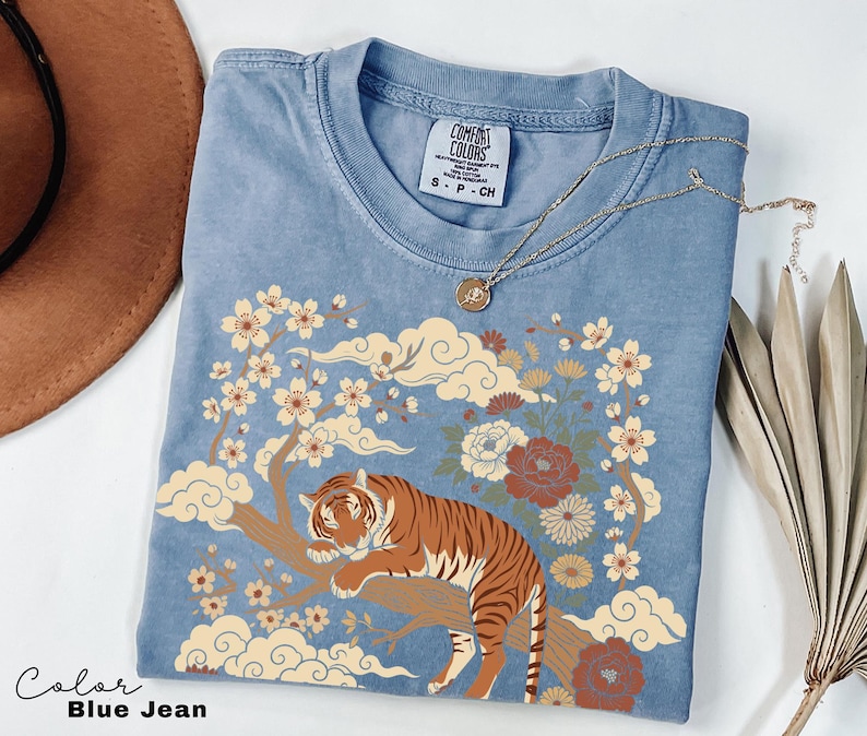 May include: Blue jean colored t-shirt with a tiger graphic. The tiger is resting on a tree branch with flowers and clouds. The shirt has a crew neck and short sleeves. A gold necklace is draped across the shirt.