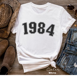 May include: White t-shirt with the year 1984 printed in black block letters.