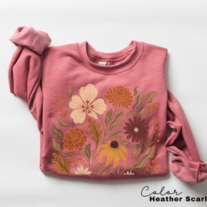 Puede incluir: Una sudadera de cuello redondo en color rojo escarlata jaspeado con un estampado gr&aacute;fico de varias flores en tonos crema, naranja, amarillo y marr&oacute;n oscuro con follaje verde. El texto "Color Heather Scarlet Red" aparece abajo a la derecha.