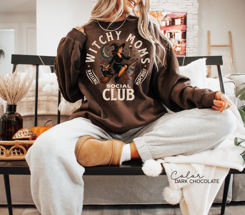 May include: Dark chocolate-coloured sweatshirt featuring the text "Witchy Moms Social Club" and an illustration of a witch on a broom, holding a glass of wine. The sweatshirt is worn with grey sweatpants.