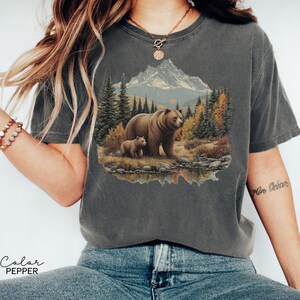 May include: Dark gray t-shirt featuring a detailed graphic of a bear and cub in a forest scene with a mountain backdrop. The image is printed on a comfortable, casual tee. The text "Color Pepper" is at the bottom.