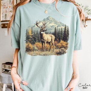 May include: A light blue t-shirt featuring a detailed illustration of a deer in a mountain landscape. The artwork depicts a deer with large antlers standing in a field with trees and a mountain range in the background. The shirt has a relaxed fit.