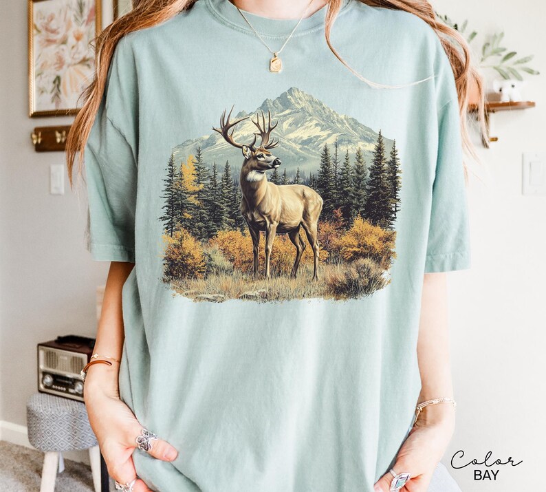 May include: A light blue t-shirt featuring a detailed illustration of a deer in a mountain landscape. The artwork depicts a deer with large antlers standing in a field with trees and a mountain range in the background. The shirt has a relaxed fit.