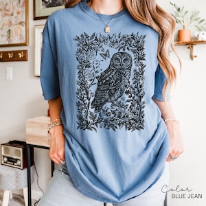 May include: A blue jean colored t-shirt featuring a detailed black and white illustration of an owl within a floral frame. The owl is the focal point, with intricate feather details. The shirt has short sleeves and a relaxed fit, suitable for casual wear.