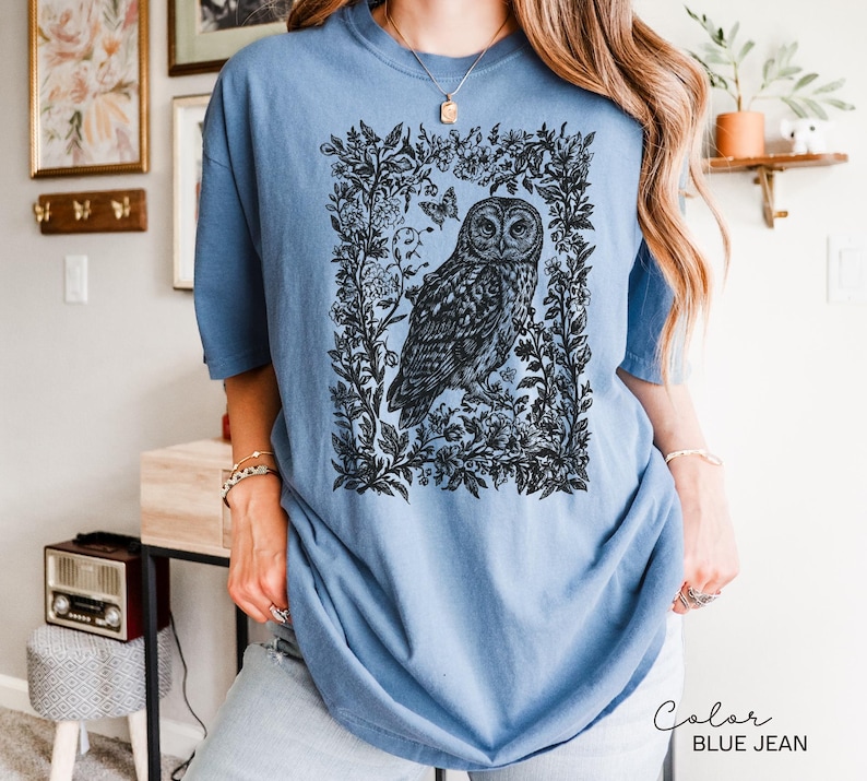 May include: A blue jean colored t-shirt featuring a detailed black and white illustration of an owl within a floral frame. The owl is the focal point, with intricate feather details. The shirt has short sleeves and a relaxed fit, suitable for casual wear.