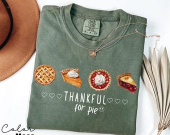 Può includere: Maglietta verde a tema Ringraziamento. Presenta quattro tipi diversi di torta con il testo "THANKFUL for pie" e un'emoticon sorridente.