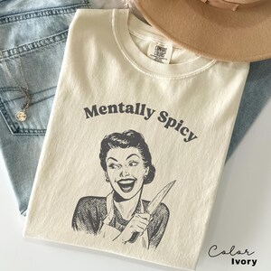 Puede incluir: Camiseta de color marfil con el texto "Mentally Spicy" y una ilustraci&oacute;n retro de una mujer sosteniendo un cuchillo.