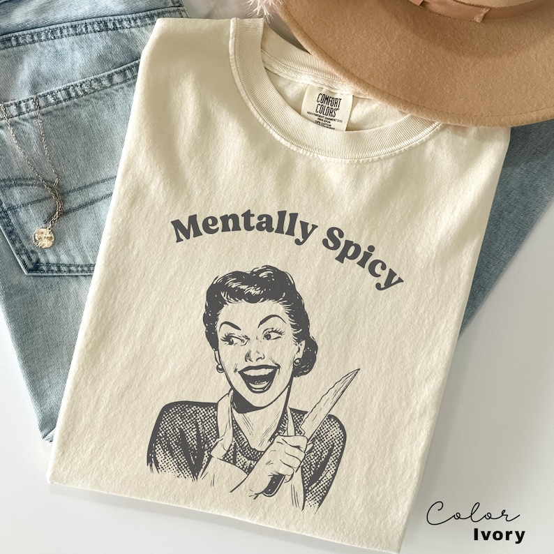 Puede incluir: Camiseta de color marfil con el texto "Mentally Spicy" y una ilustraci&oacute;n retro de una mujer sosteniendo un cuchillo.