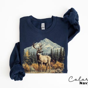 May include: Navy blue sweatshirt featuring a detailed graphic of a deer with large antlers standing in front of a mountain range and evergreen trees. The sweatshirt has a crew neck and long sleeves. The words "Color Navy" are written in cursive.