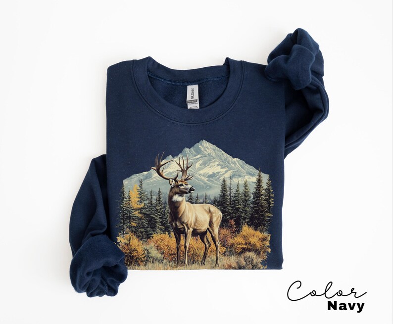 May include: Navy blue sweatshirt featuring a detailed graphic of a deer with large antlers standing in front of a mountain range and evergreen trees. The sweatshirt has a crew neck and long sleeves. The words "Color Navy" are written in cursive.