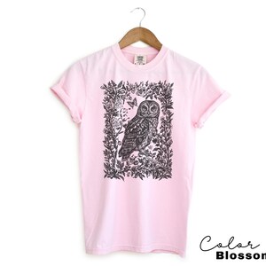 May include: A light pink t-shirt featuring a black and white illustration of an owl within a floral frame. The text "Color Blossom" is written in a cursive font at the bottom right corner. The shirt is hanging on a wooden hanger.