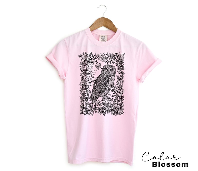 May include: A light pink t-shirt featuring a black and white illustration of an owl within a floral frame. The text "Color Blossom" is written in a cursive font at the bottom right corner. The shirt is hanging on a wooden hanger.