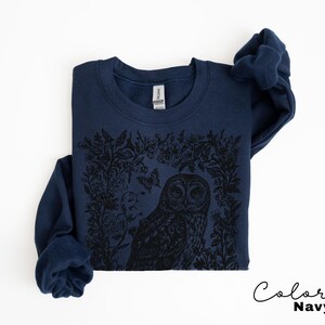 May include: Navy blue sweatshirt featuring a black graphic of an owl surrounded by floral and butterfly elements. The sweatshirt has a crew neck and long sleeves. The text "Color Navy" is written in cursive in the bottom right corner.