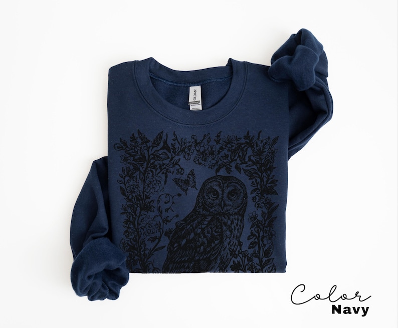 May include: Navy blue sweatshirt featuring a black graphic of an owl surrounded by floral and butterfly elements. The sweatshirt has a crew neck and long sleeves. The text "Color Navy" is written in cursive in the bottom right corner.