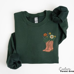 Puede incluir: Sudadera verde bosque con un gr&aacute;fico de botas de vaquero color melocot&oacute;n llenas de flores coloridas. El texto "Color Forest Green" est&aacute; en la esquina inferior derecha. La sudadera est&aacute; doblada.