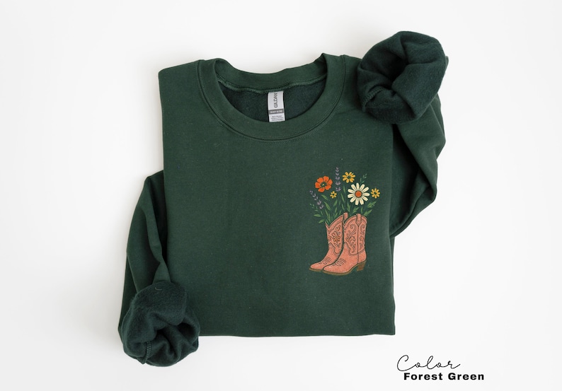 Puede incluir: Sudadera verde bosque con un gr&aacute;fico de botas de vaquero color melocot&oacute;n llenas de flores coloridas. El texto "Color Forest Green" est&aacute; en la esquina inferior derecha. La sudadera est&aacute; doblada.