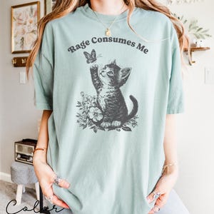 May include: A light blue t-shirt with a black and white illustration of a cat reaching for a butterfly. The text "Rage Consumes Me" is printed in black on the shirt.