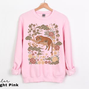 May include: Light pink sweatshirt featuring a detailed illustration of a tiger resting on a tree branch, surrounded by flowers and clouds. The text "Color Light Pink" is at the bottom. The design is in shades of brown, gold, and pink.
