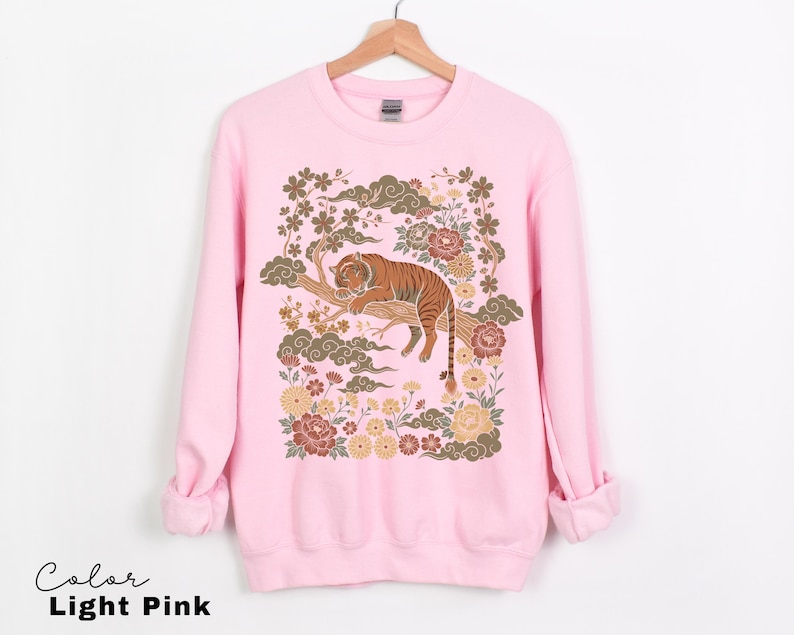 May include: Light pink sweatshirt featuring a detailed illustration of a tiger resting on a tree branch, surrounded by flowers and clouds. The text "Color Light Pink" is at the bottom. The design is in shades of brown, gold, and pink.