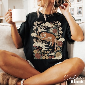 May include: Black t-shirt featuring a tiger resting on a tree branch, surrounded by floral and cloud designs. The shirt has the text "Color Black" in a script font. The design includes shades of brown, orange, cream, and burgundy.