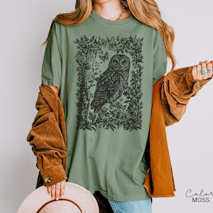 May include: Sage green t-shirt featuring a detailed black illustration of an owl within a floral frame. The design includes butterflies and foliage. The shirt is a relaxed fit.