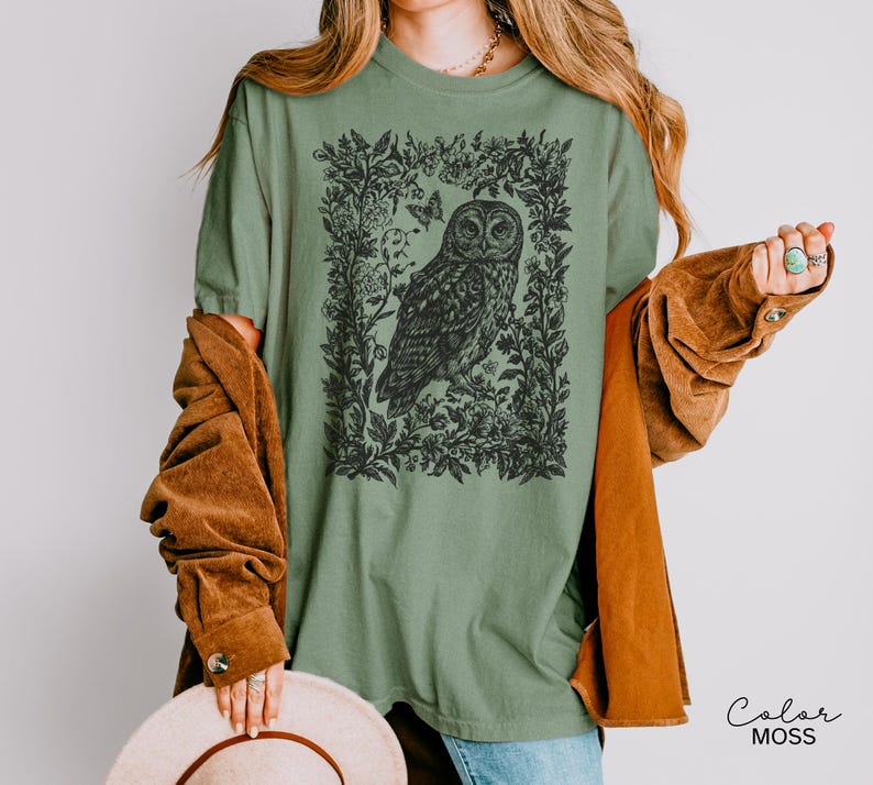 May include: Sage green t-shirt featuring a detailed black illustration of an owl within a floral frame. The design includes butterflies and foliage. The shirt is a relaxed fit.