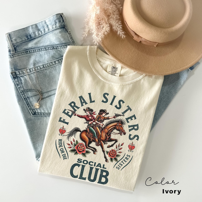 May include: Ivory t-shirt with a graphic of two cowgirls riding a horse. The graphic is surrounded by a circle with the text "Feral Sisters Social Club" and the words "Ride or Die" and "Sisters".