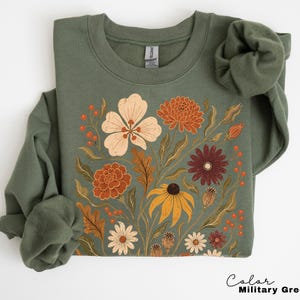 Puede incluir: Una sudadera de cuello redondo en color verde militar con un estampado gr&aacute;fico central de diversas flores y follaje en tonos crema, naranja, &oacute;xido y rojo oscuro. La prenda de manga larga presenta un cuello y pu&ntilde;os acanalados.
