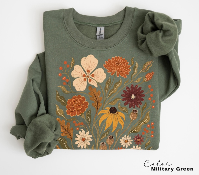 Puede incluir: Una sudadera de cuello redondo en color verde militar con un estampado gr&aacute;fico central de diversas flores y follaje en tonos crema, naranja, &oacute;xido y rojo oscuro. La prenda de manga larga presenta un cuello y pu&ntilde;os acanalados.