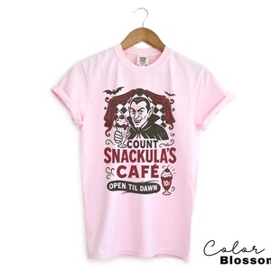 May include: A light pink t-shirt featuring a graphic of Count Dracula holding a milkshake, with the text "Count Snackula's Cafe" and "Open Til Dawn". The design includes bats and a checkered background.