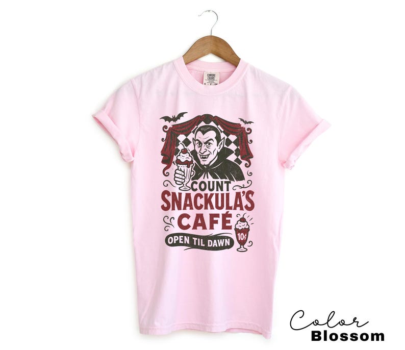 May include: A light pink t-shirt featuring a graphic of Count Dracula holding a milkshake, with the text "Count Snackula's Cafe" and "Open Til Dawn". The design includes bats and a checkered background.