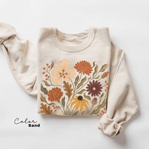 Puede incluir: Una sudadera de cuello redondo color arena con un estampado gr&aacute;fico de varias flores y follaje en tonos naranja, amarillo, marr&oacute;n y verde apagado. El texto "Color Sand" est&aacute; impreso en negro en la esquina inferior izquierda de la imagen.
