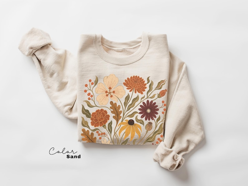 Puede incluir: Una sudadera de cuello redondo color arena con un estampado gr&aacute;fico de varias flores y follaje en tonos naranja, amarillo, marr&oacute;n y verde apagado. El texto "Color Sand" est&aacute; impreso en negro en la esquina inferior izquierda de la imagen.