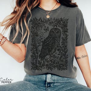 May include: Pepper-colored t-shirt featuring a detailed black illustration of an owl surrounded by floral elements and butterflies. The design is centered on the shirt, creating a vintage aesthetic. The shirt's color is labeled as "Pepper".
