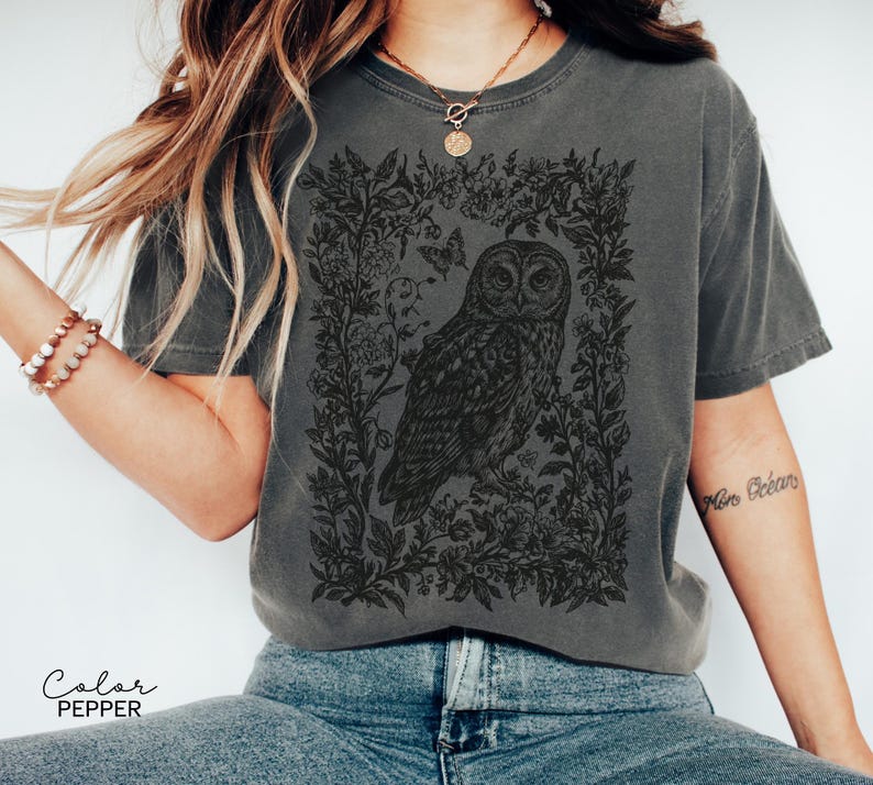 May include: Pepper-colored t-shirt featuring a detailed black illustration of an owl surrounded by floral elements and butterflies. The design is centered on the shirt, creating a vintage aesthetic. The shirt's color is labeled as "Pepper".