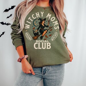 May include: Military green sweatshirt with a circular graphic featuring a witch riding a broom, a black cat, and the text "Witchy Moms Social Club." The design includes a crescent moon and stars. The words "Raising" and "Mischief" are also included.