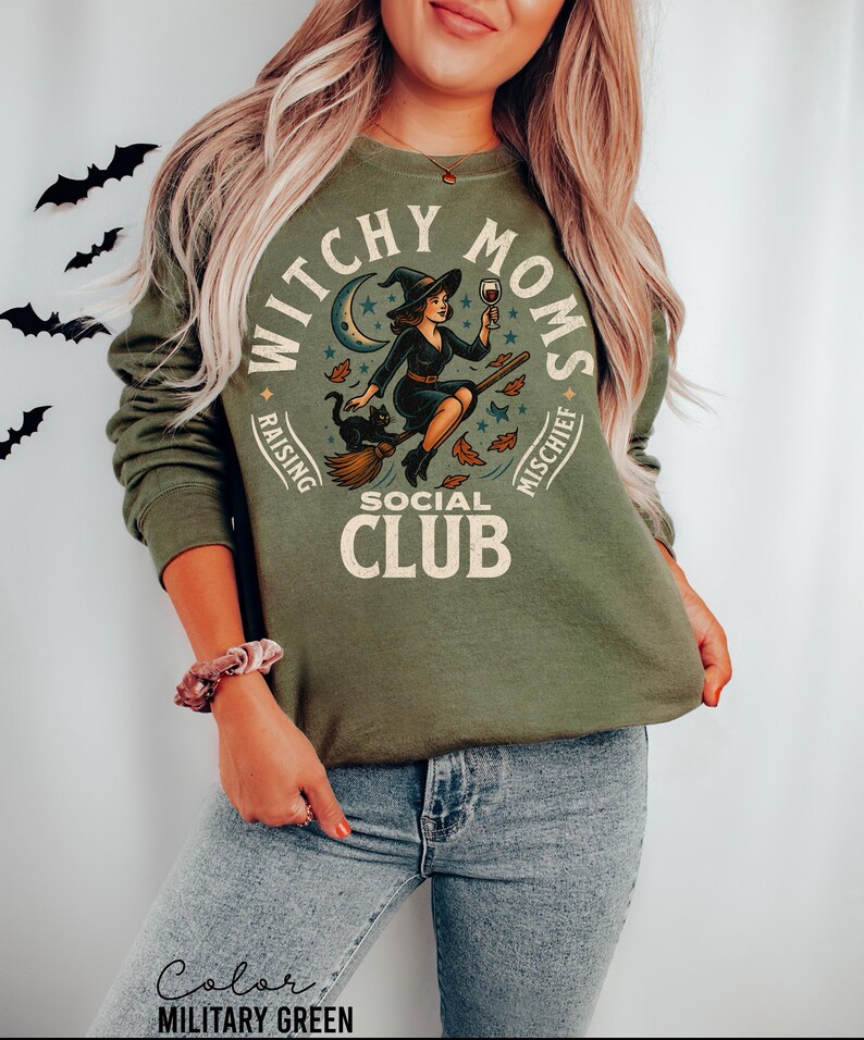 May include: Military green sweatshirt with a circular graphic featuring a witch riding a broom, a black cat, and the text "Witchy Moms Social Club." The design includes a crescent moon and stars. The words "Raising" and "Mischief" are also included.