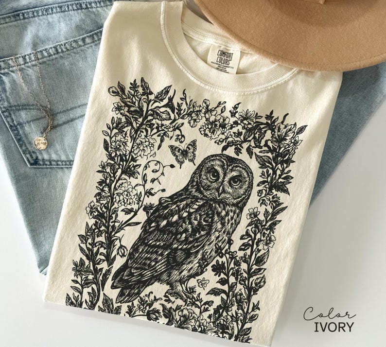 May include: Ivory-colored t-shirt featuring a detailed black and white illustration of an owl surrounded by floral elements and a butterfly. The shirt is folded, with a hat and jeans in the background.