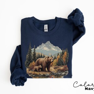 May include: Navy blue sweatshirt featuring a detailed illustration of a bear and cub in a forest setting with mountains in the background. The artwork includes trees, a lake, and autumn colors. The text "Color Navy" is written in cursive.