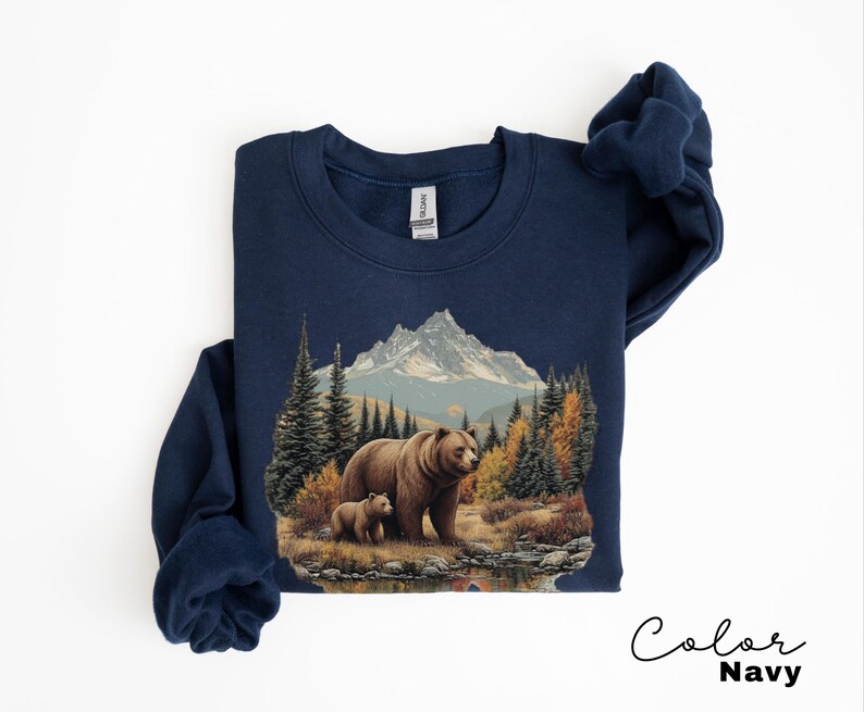 May include: Navy blue sweatshirt featuring a detailed illustration of a bear and cub in a forest setting with mountains in the background. The artwork includes trees, a lake, and autumn colors. The text "Color Navy" is written in cursive.