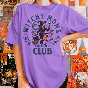 Puede incluir: Una camiseta de color lila con un dise&ntilde;o circular que dice "Witchy Moms Social Club". El dise&ntilde;o muestra una bruja en una escoba con un gato negro, una luna creciente y estrellas. Las palabras "Raising" y "Mischief" tambi&eacute;n est&aacute;n incluidas.
