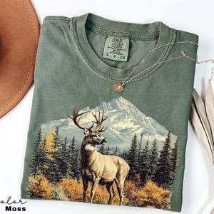 May include: An olive green t-shirt featuring a mountain scene with a deer in the foreground. The graphic includes a mountain range, trees, and autumn foliage. The shirt has a crew neck and a small tag on the collar.