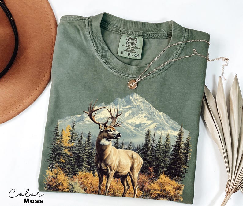 May include: An olive green t-shirt featuring a mountain scene with a deer in the foreground. The graphic includes a mountain range, trees, and autumn foliage. The shirt has a crew neck and a small tag on the collar.