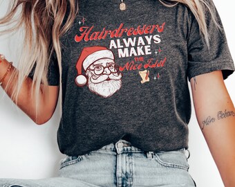 Op de afbeelding: Een donkergrijs T-shirt met een rode en witte illustratie van de kerstman. De tekst "Hairdressers Always Make the Nice List" staat op het T-shirt gedrukt.