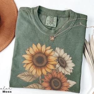 May include: A green t-shirt featuring a sunflower design. The shirt has a distressed look and is made from a soft, comfortable fabric. The text "COMFORT COLORS" is visible on the tag.